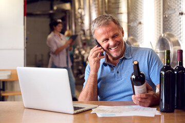 Mature Man On Mobile Phone With Laptop Checking Label On Bottle Inside Winery 