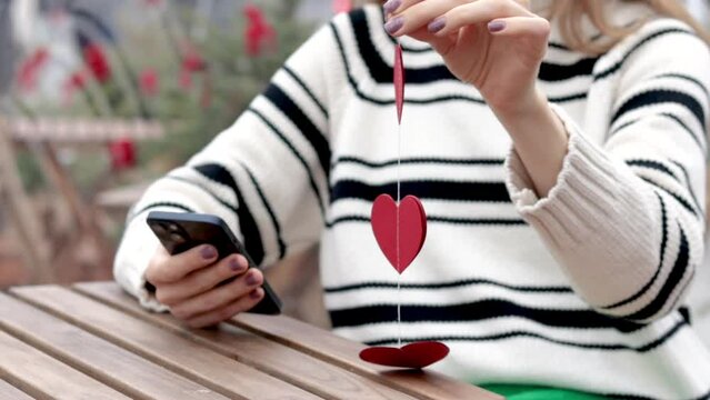 woman sitting in on table with paper hearts garland in one hand using smartphone with another,girl hugging red balloon heart shape.celebrate love valentine day coffee shop inside transparent balloon - Powered by Adobe