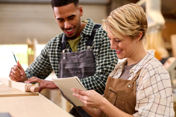 Male And Female Apprentices Working As Carpenters In Furniture Workshop Measure Wood And Take Notes
