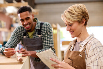 Male And Female Apprentices Working As Carpenters In Furniture Workshop Measure Wood And Take Notes