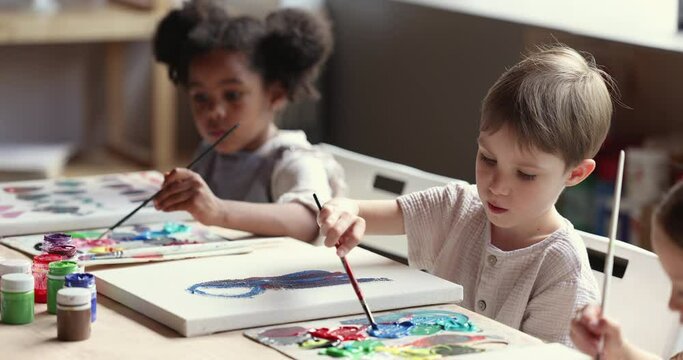 Multiracial kids, little painters engaged in painting art class in studio. Diverse children sit at table hold paintbrush drawing on canvas, looking interested and inspired. Art therapy, creative hobby