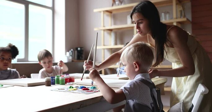 Young artist woman, teacher lead art class for little multiethnic preschooler kids seated at table holding paintbrushes and painting pictures look interested take part in creative day-care art studio