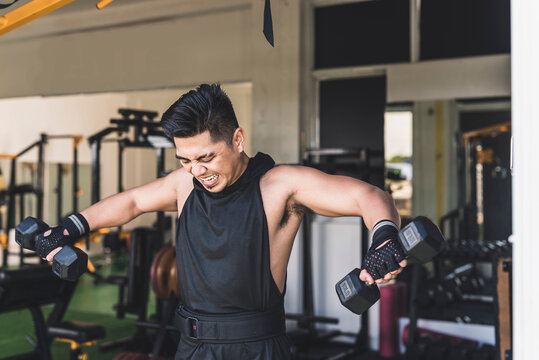 An asian guy strugling to do a set of standing dumbbell side lateral raises at the gym. Enduring the last reps, feeling muscle burn. Weight training at the gym.