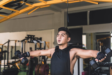 A tanned and handsome asian guy doing standing dumbbell side lateral raises at the gym. Wearing a black hooded low cut tank top. Weight training at the gym.