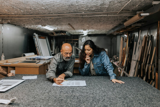 Mature bald owner discussing blueprint with female client at workbench