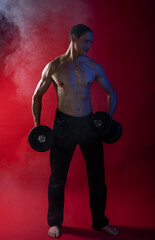 bare-chested man in colored light. showing muscles posing with dumbbells and smoke. standing or on the floor. red background. Studio