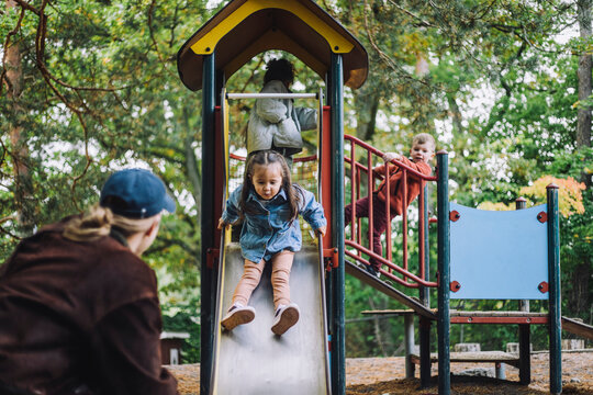Male teacher looking at girl playing on slide in playground