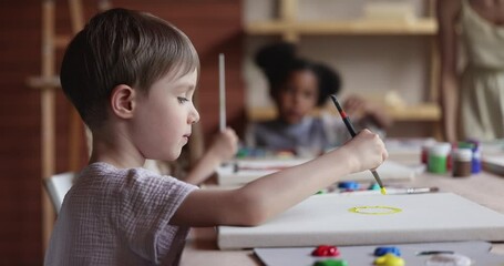 Diverse kids gather in art class lead by female teacher, to develop creative ability, gain new skill, enjoy hobby, sit together at table, hold brushes painting pictures on canvas in artistic workshop