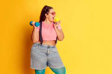 Portrait of overweight young woman posing with dumbbell and apple over yellow studio background. Diet and sport combination