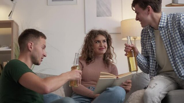 Different interests. Happy friends. Home leisure. Inspired two men cheering with beer while blaming woman reading book on sofa light room interior.