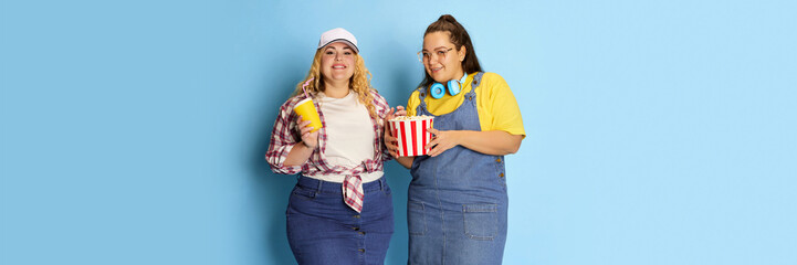 Portrait of two fat, beautiful women, friends posing with popcorn basket over blue studio background. Banner