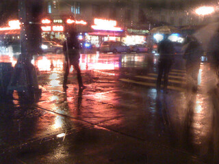 Shadowed figures carrying umbrellas walk down a busy city street on a rainy night.