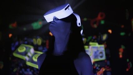 Young woman in VR headset looking around to abstract cyber world and neon social media icons. Girl wearing virtual or augmented reality 3D glasses at glowing background. Immersing in metaverse - Powered by Adobe