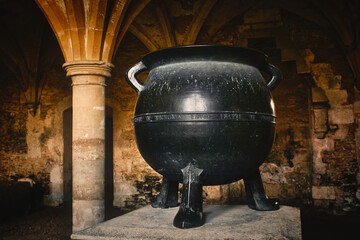 A big black cast iron metal cauldron in a medieval castle basement under an arch