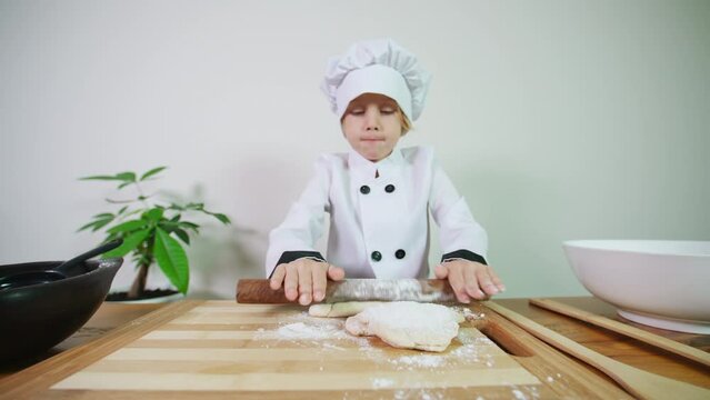 Sliding view of a cute 5 years old caucasian boy dressed like a chef rolling out pastry with a rolling pin preparing it for baking a pie, pizza or biscuits in the oven. A boy helps in the kitchen