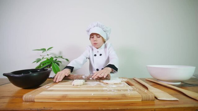 A cute 5 years old caucasian boy dressed like a chef and focused on rolling out pastry with a rolling pin preparing it for baking pizza or biscuits in oven. A boy helps in the kitchen