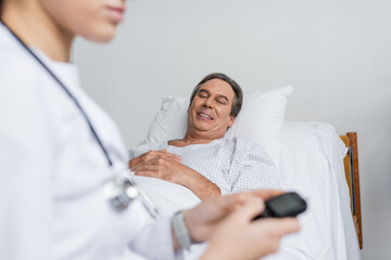 Smiling elderly patient looking at blurred doctor with glucometer in hospital ward.