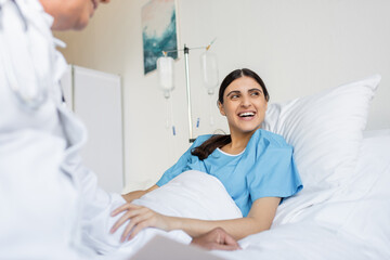 Obraz premium Cheerful patient in gown lying on bed near blurred doctor in clinic.