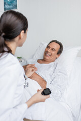 Fototapeta premium Smiling senior patient looking at blurred doctor with glucometer in clinic.