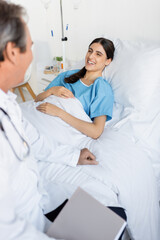 Obraz premium Cheerful patient looking at blurred senior doctor while lying on bed in hospital ward.