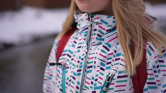 Unrecognizable teen girl zipping coat leaving on winter day outdoors. Blond Caucasian teenager with long hair in warm clothing walking away at river shore. Weather and lifestyle