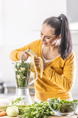 A woman in the kitchen cuts a banana in a blender together with spinach, makes a healthy smoothie