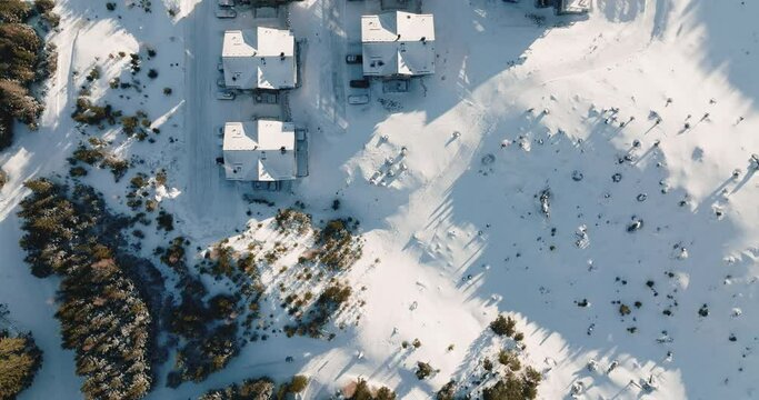Drone flies directly above snow covered winter ski resort village buildings, chalet countryside on sunny morning.