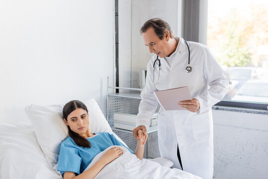 Senior Doctor With Paper Folder Holding Hand Of Brunette Patient In Hospital Ward.