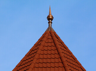 pointy spire on turret with hexagonal sloped building roof top. red clay tile roof. copper spike or spire at the tip. ridge tiles. blue sky background. building materials and construction concept. © Istvan