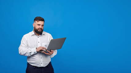 Naklejka na ściany i meble A bearded man holds a laptop in his hands on a blue background. A solid man in a white shirt works at a laptop and looks into the camera.