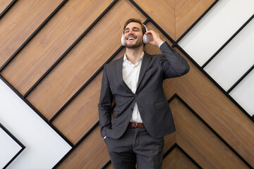 a young businessman in a strict gray suit enjoys listening to music on headphones against the background of an office wall