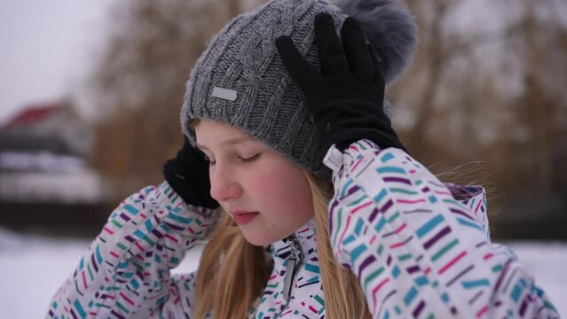 Close-up side view teenage cute girl adjusting hat turning looking at camera smiling in slow motion. Portrait of confident pretty Caucasian teenager posing outdoors on cold winter day in park