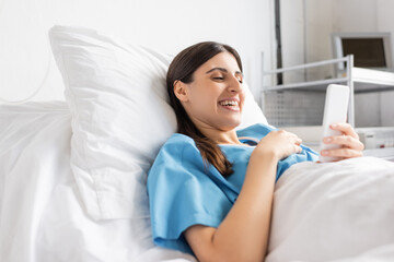 Cheerful patient in gown using smartphone while lying on bed in clinic.