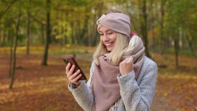 Side view of attractive, blonde woman walking in park in autumn. Beautiful, young female using smartphone, texting, showing hurray, smiling. Concept of sincere emotions.