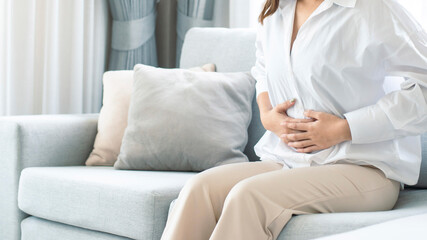 Young woman wearing casual clothes suffering from menstrual pain, feeling sick to her stomach, holding belly, having abdominal cramps during period and lying down on sofa in the living room at home. - obrazy, fototapety, plakaty