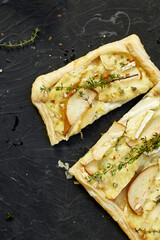 Pie baked in puff pastry with gorgonzola cheese and pears on black table, cutting pizza with pear, thyme, copy space, top view.