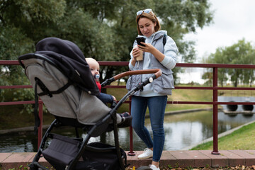 young mother with a baby stroller on a walk in the park with a phone in her hands