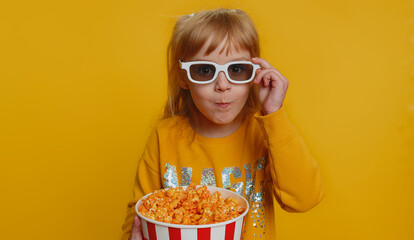 Excited young toddler school girl in 3D glasses eating popcorn, watching interesting tv serial,...