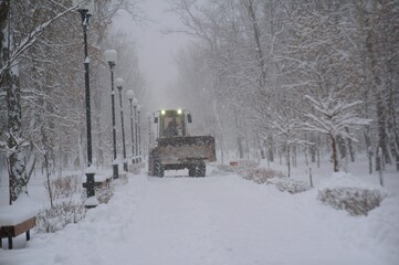 A snowplow removes snow in a city park. It's snowing.