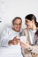 Positive patient pointing at smartphone near daughter in hospital ward.
