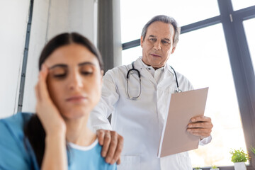 Senior doctor with paper folder calming blurred patient in hospital ward.