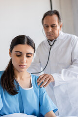 patient in gown sitting near blurred doctor with stethoscope in clinic.