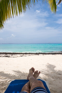 The feet of a person resting on a deck chair under a palm tree, on a paradisiacal beach in the Caribbean