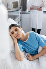 Displeased woman in patient gown lying on bed near blurred doctor in hospital ward.