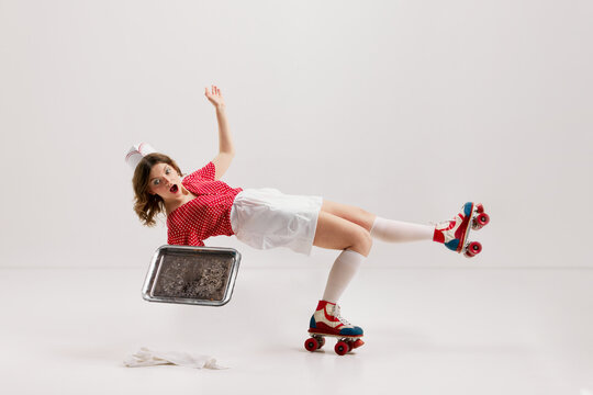 Emotional excited young girl, stylish retro waitress in american fashion style of 70s, 80s uniform rollerblade over gray background. Falling