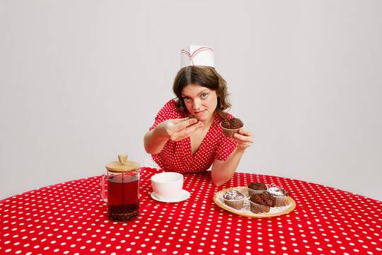 Stylish young girl in retro american fashion style of 70s, 80s uniform sitting at table and tasting cupcakes with coffee over grey background. Pop art Photography - Powered by Adobe