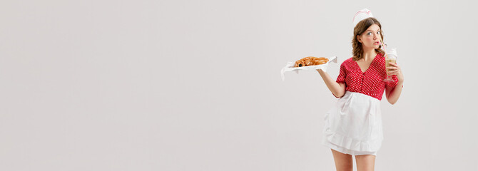 Portrait of young pretty waitress, woman in retro american fashion style of 70s, 80s uniform treats with bun over grey background. Banner