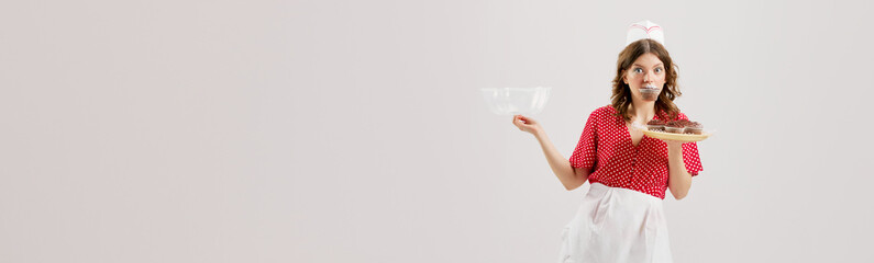 Portrait of young female waitress, woman in retro american fashion style of 70s, 80s uniform treats with chocolate cupcakes over grey background. Banner - Powered by Adobe