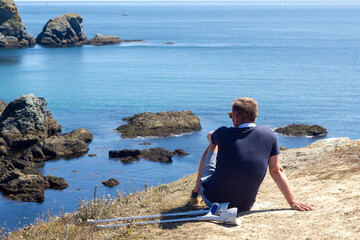 Unrecognizable disabled man sitting with his crutches alongside on the rock admiring a beautiful sea scenery of Belle-Ile-En-Mer, Brittany, France