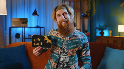 Hippie redhead man at home couch showing pointing on gift discount certificate voucher coupon for store holidays sale. Young hipster guy with shopping christmas surprise card in night evening room